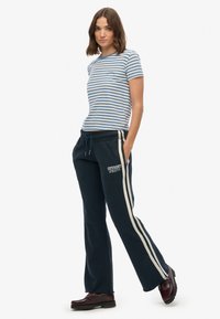 Striped blue and white t-shirt, fitted; navy sweatpants with white side stripes and "SUPERDRY ATHLETIC" text, worn with dark shoes.