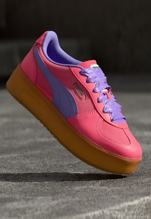 Jasnoróżowy but sportowy Puma z fioletowymi sznurowadłami i logo oraz grubą żółtą podeszwą, na rozmytym szarym tle.