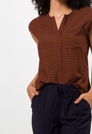 Kvinde iført ærmeløs brun og sort zigzag-mønstret top, der er stoppet ned i løse sorte bukser med hænderne i lommerne.