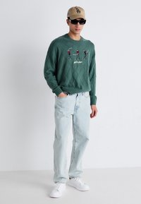 Groene trui met ruitpatroon en geborduurde golffiguren, lichtblauwe jeans, witte sneakers en een beige pet. Zonnebril maakt de look compleet.