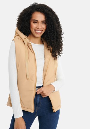 Beige gewatteerde vest met een capuchon, rits aan de voorkant, trekkoord en zijzakken, gedragen over een witte langemouwenshirt en blauwe spijkerbroek.