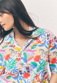 Kurzärmlige Bluse aus leichtem Stoff mit einem bunten tropischen Druck, der Blumen, Tukane, Zebras und lebendige Blätter zeigt.