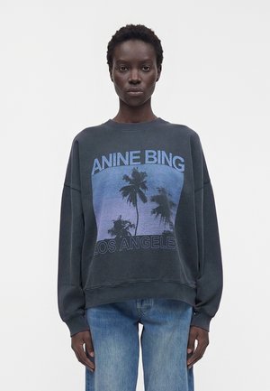 Personne à la peau foncée portant un sweat-shirt noir oversize avec un motif de palmier bleu et le texte « ANINE BING LOS ANGELES », associé à un jean bleu.