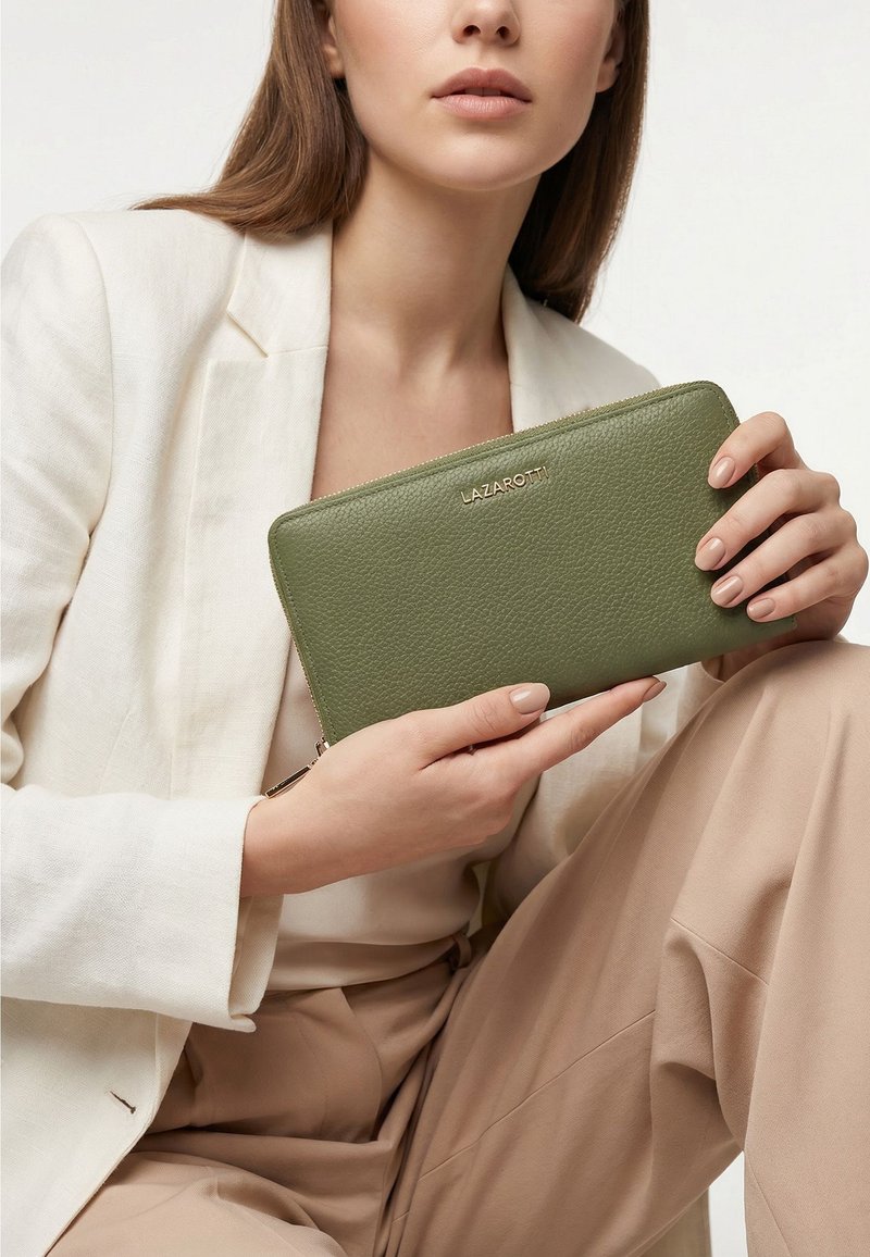 Mujer con conjunto beige sostiene una cartera con cremallera Lazarotti de color verde oliva con cuero texturizado y logo dorado en el frente.