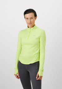 Vrouw in een limoen groene sporttop met lange mouwen en kwart-rits, gecombineerd met donkergrijze leggings, staande tegen een effen witte achtergrond.
