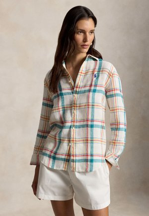 Žena oblečená v košeli s dlhým rukávom a károvaným vzorom v odtieňoch tyrkysovej, červenej a žltej, a v bielych šortkách, stojaca pred jednoduchým pozadím.