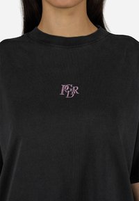 T-shirt de algodão preto com decote redondo e mangas curtas. Pequeno logótipo bordado em rosa claro no peito. Textura suave.