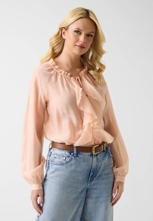 Femme blonde portant un chemisier rose clair à volants, rentré dans un jean bleu clair taille haute avec une ceinture marron, debout devant un fond uni.