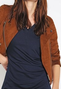 Veste en daim marron au design ajusté, avec fermeture éclair à l'avant et coutures détaillées, portée sur un t-shirt col en V bleu foncé.