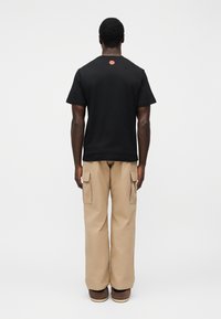 Μαύρο κοντομάνικο μπλουζάκι με στρογγυλή λαιμόκοψη και μικρή κόκκινη λεπτομέρεια στο πίσω μέρος, σε συνδυασμό με μπεζ παντελόνι cargo με πλαϊνές τσέπες.