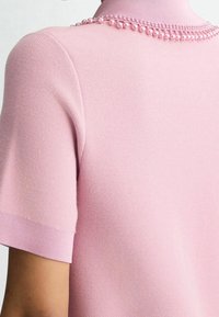 Polo à manches courtes, rose clair, en tissu tricoté texturé, avec un col haut orné de perles roses décoratives le long de l'encolure.