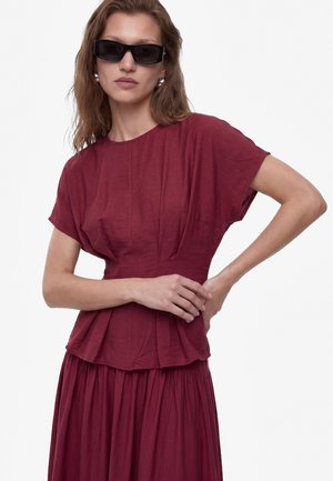 Vrouw draagt een donkerrode jurk met korte mouwen met geplooide taille en rok, gecombineerd met zwarte rechthoekige zonnebril en zilveren oorbellen.