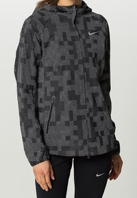 Veste Nike à motifs noir et blanc avec fermeture éclair à l'avant, capuche et poches latérales ; fabriquée dans un tissu léger et texturé.