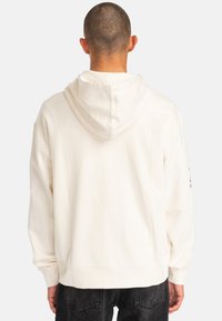 Cream-colored hoodie with a drawstring hood, loose fit, ribbed hem, and cuffs; features a logo on the right sleeve. Textured cotton fabric.