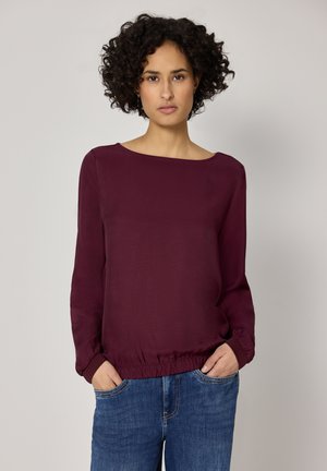 Vrouw met krullend haar, gekleed in een donkerpaarse blouse met lange mouwen en een blauwe spijkerbroek, met handen in de zakken, tegen een effen achtergrond.
