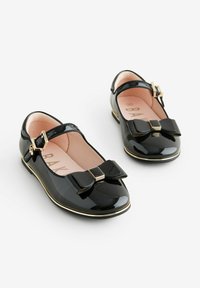 Zapatos Mary Jane de charol negro con punta cuadrada, acentos dorados y un prominente detalle de lazo en la parte delantera. Incluye correa ajustable.