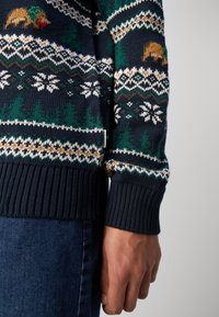 Gros plan d'une personne portant un pull bleu marine avec des sapins verts, des flocons de neige blancs et des ours bruns tricotés dans un motif.