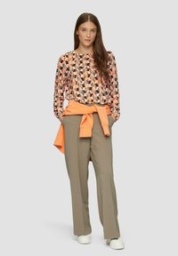 Langarmshirt met een geometrisch patroon in perzik, zwart en beige. Gecombineerd met lichtbruine wijde broek en een oranje trui om de taille geknoopt.