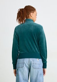 Pessoa com um rabo de cavalo trançado, vestindo uma jaqueta de veludo teal e calças cargo de cintura alta em azul claro, de costas contra um fundo simples.