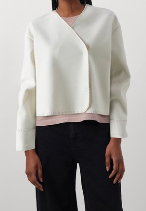 Personne portant une veste asymétrique blanche sur un haut rose clair et un pantalon noir sur un fond clair uni.