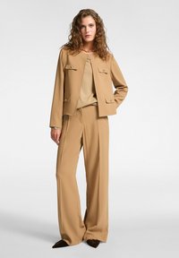 Beige sestavené sako se dvěma předními kapsami a kulatým výstřihem, spárované s odpovídajícími širokými kalhotami a béžovým topem pod ním.