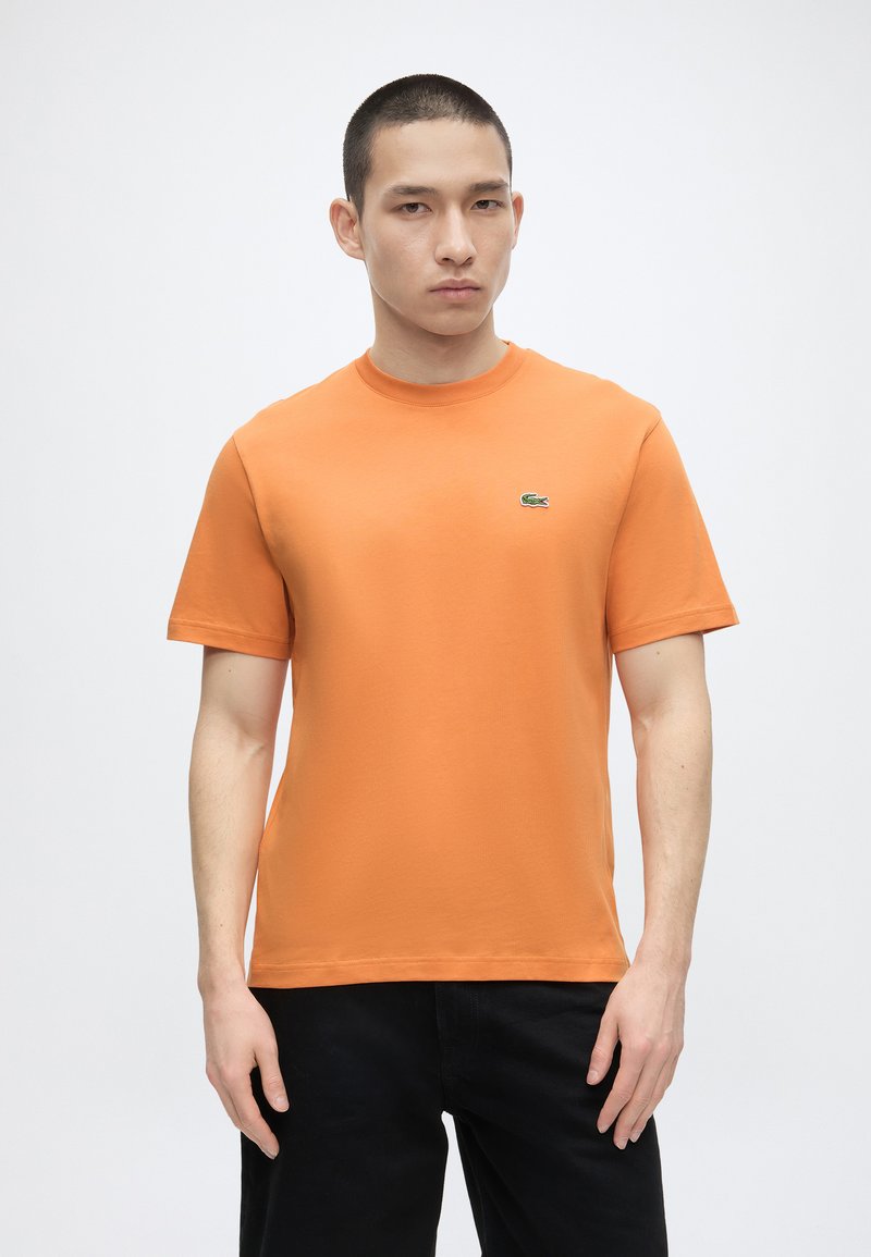 Νέος άνδρας με απλό πορτοκαλί κοντομάνικο t-shirt με μικρό πράσινο λογότυπο κροκόδειλου στο στήθος, μαύρο παντελόνι, στέκεται μπροστά σε λευκό φόντο.