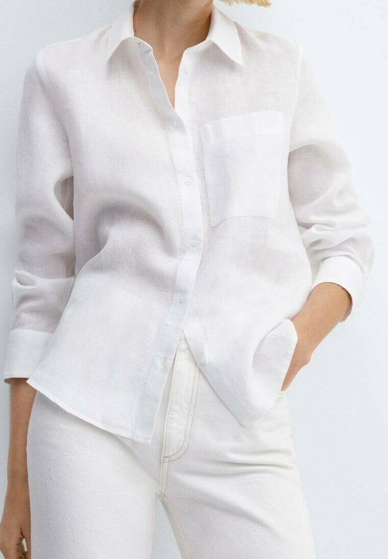 Mujer vistiendo una camisa blanca semi-translúcida de botones con un bolsillo en el pecho sobre pantalones blancos de cintura alta, con la mano en el bolsillo.