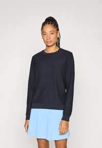 Tengerészkék, texturált pulóver kerek nyakkal és bordázott mandzsettákkal, apró logóval a mellrészen. Világoskék szoknyával párosítva.