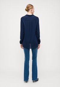 Chemise à manches longues bleu marine avec col, dotée de poignets boutonnés et d'un ourlet arrondi. Portée avec un jean flare bleu clair et des chaussures noires.