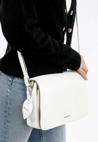 Hvid læder crossbody taske med tekstureret overflade, flaplukning og hjerteformet charm-accent. Justerbar skulderrem og sølvhardware.