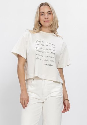 Vrouw met schouderlang blond haar die een los wit Calvin Klein T-shirt en bijpassende witte broek draagt, staand tegen een effen achtergrond.