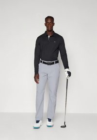 Polo preta de manga comprida, calças de golfe cinzentas, cinto preto, sapatos brancos com detalhes azuis, segurando um taco de golfe, em pé contra um fundo simples.