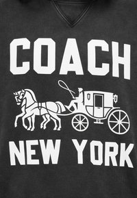 Μαύρο ύφασμα με λευκό κείμενο που γράφει "COACH NEW YORK" και ένα σχέδιο με άμαξα που σύρεται από άλογο, όπου ο οδηγός κρατά τα χαλινάρια.