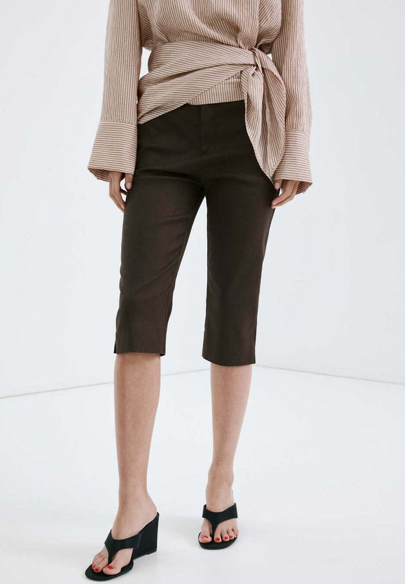 Femme portant un pantalon marron foncé jusqu'aux genoux, un chemisier beige rayé à manches longues noué à la taille, et des sandales compensées noires sur un sol blanc.