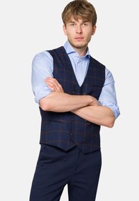 Granatowa kamizelka w kratę z brązowymi akcentami na jasnoniebieskiej koszuli. Materiał wygląda na teksturowany; ma dekolt w kształcie litery V i dwie przednie kieszenie.