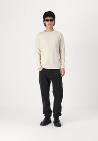 Chemise à manches longues texturée crème avec des manches raglan, jean noir ample, et chaussures à lacets noires, portées avec des lunettes de soleil sombres.