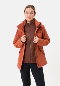 Giacca arancione impermeabile con cappuccio, indossata sopra un fleece marrone con zip. Include chiusura con zip, tasche laterali e un design aderente.