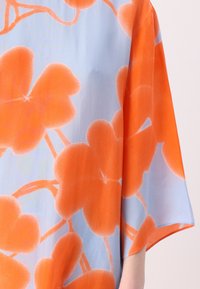 Trumpa rankovėmis blusa, pagaminta iš lengvos medžiagos, su dideliais oranžiniais gėlių raštais ant minkšto mėlyno fono. Lygus audinys ir laisva siluetas.