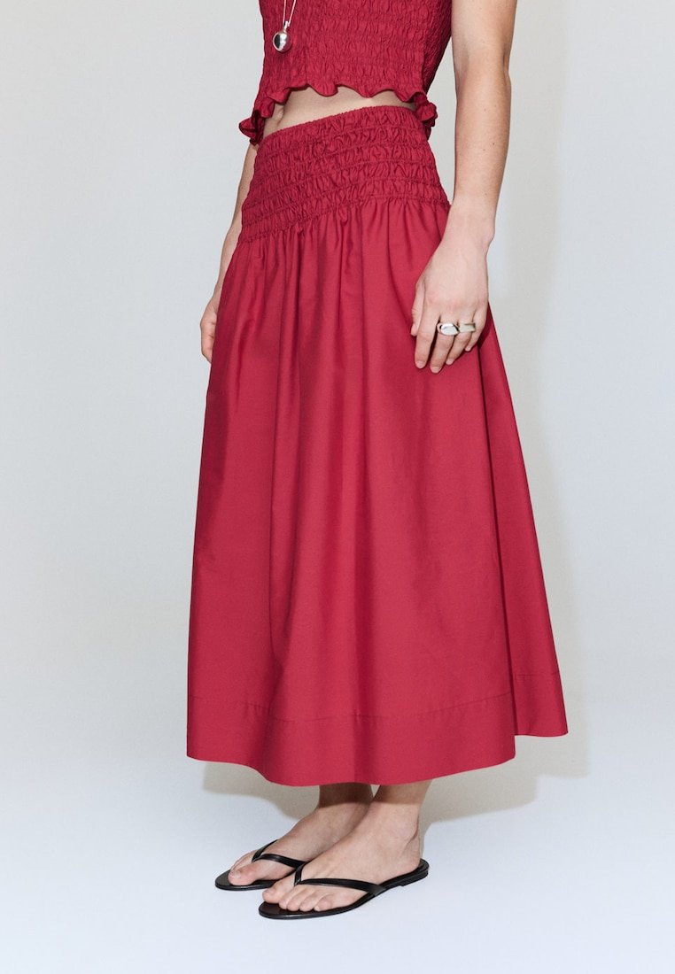 Femme portant une jupe midi rouge taille haute avec une ceinture smockée, un top court rouge, des tongs noires et des bagues argentées à la main droite.