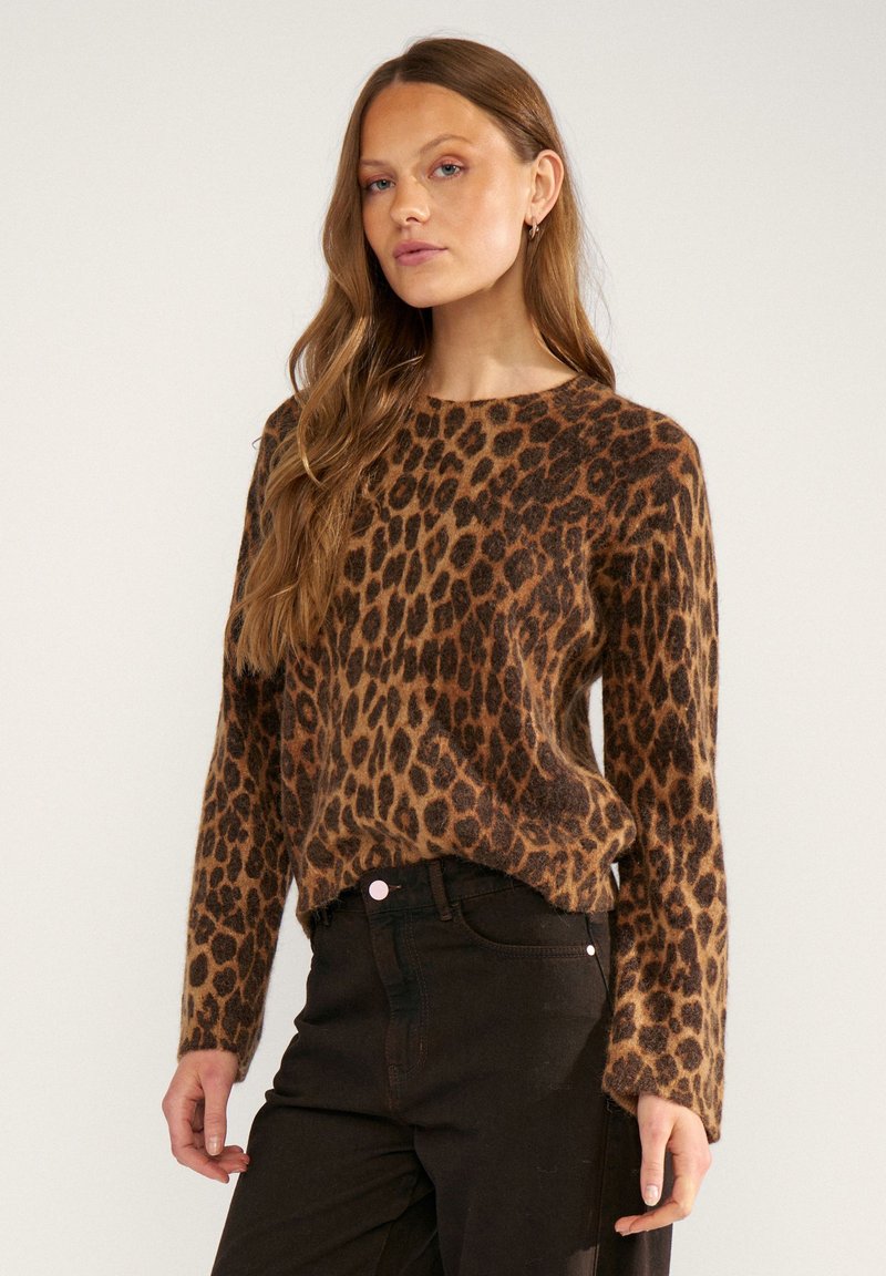 Femme aux cheveux longs portant un pull imprimé léopard marron et noir et un pantalon noir sur un fond clair uni.