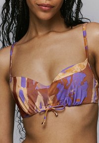 Bikini-top met een mix van paarse en bruine abstracte patronen, met dunne banden en een strikdetail in het midden. Zacht, gestructureerd materiaal.