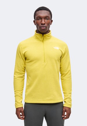 Homme portant une polaire zippée jaune vif texturée avec un col montant, debout devant un fond clair uni.