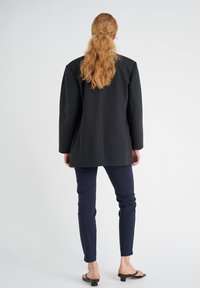 Černý oversized blejzr s ostrými rameny, spárovaný s úzkými námořnickými kalhotami a černými skluznými sandály. Hladká tkanina; minimalistický design.