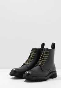 Bottes de cheville en cuir noir avec des lacets verts, toe rond et semelle en caoutchouc épaisse. Comprend des languettes en haut pour un enfilage facile.
