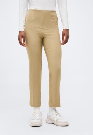 Άτομο που φοράει μπεζ παντελόνι στενής εφαρμογής, λευκή μακρυμάνικη μπλούζα, λευκές κάλτσες και λευκά αθλητικά παπούτσια, στέκεται σε απλό φόντο.