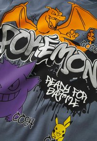 Grå tygskjorta med färgglada Pokémon-grafiker: Charizard i orange, Gengar i lila och Pikachu i gult med fet text som säger "REDO FÖR STRID."