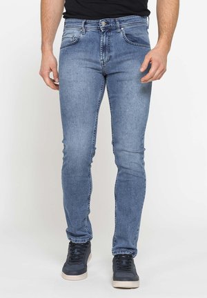 Uomo che indossa jeans blu slim fit e sneakers nere, in piedi con le mani rilassate lungo i fianchi davanti a uno sfondo neutro.