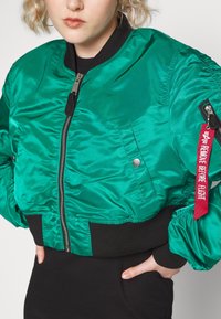 Groene satijnen bomberjack met een zwarte geribbelde tailleband en manchetten, ritssluiting aan de voorkant en een rode tag met de tekst "VERWIJDER VOOR VLUCHT."