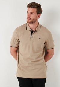 Beige pólóing fekete gallérral és ujjtövénnyel, három gombbal és egy kis logóval a mellkasán. Texturált pamut anyag.