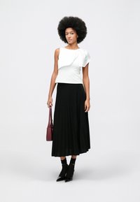 Woman with afro hairstyle wearing a white ruffled sleeveless top, black pleated midi skirt, black socks with heels, and holding a burgundy bag.